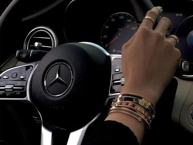 Close-up of a hand wearing gold bracelets holding the steering wheel of a Mercedes-Benz car.
