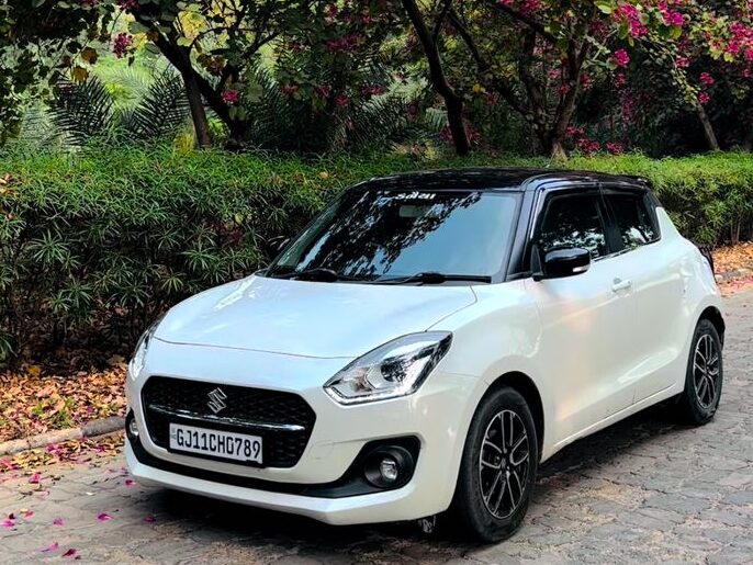 Front-side view of a white Maruti Suzuki Swift parked on a tree-lined street.